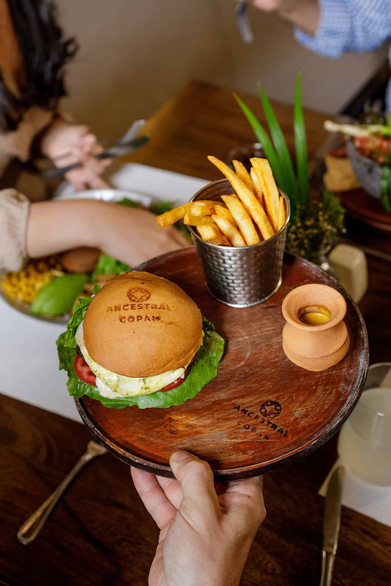 Taste the Local Difference Signature Ancestral Copán burger with branded bun, golden fries, and traditional clay sauce pot served on wooden board at Balam Bistro
