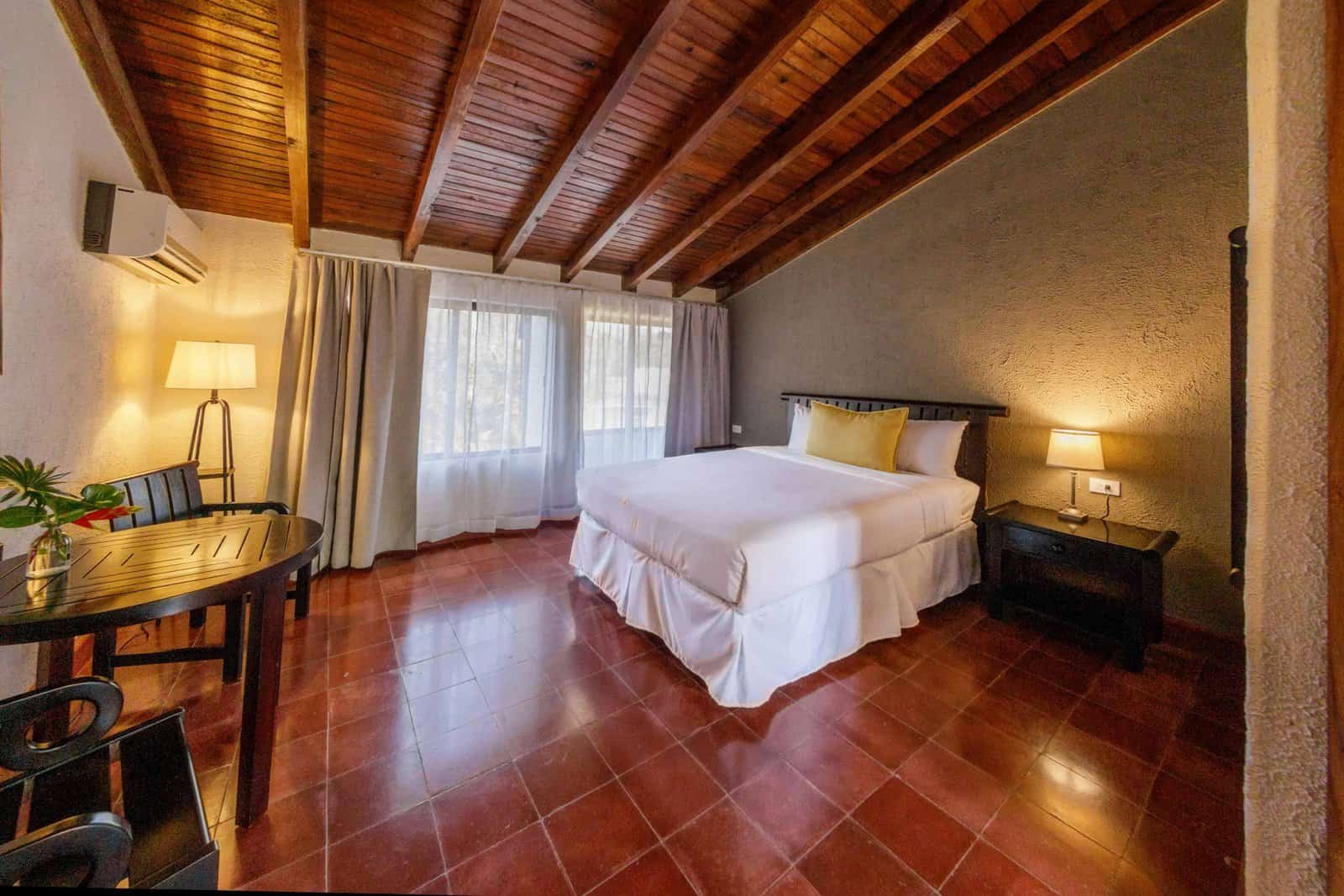 Premium king bedroom with exposed wooden beam ceiling, terra cotta floors, warm lighting, and sitting area at Ancestral Copán Hotel